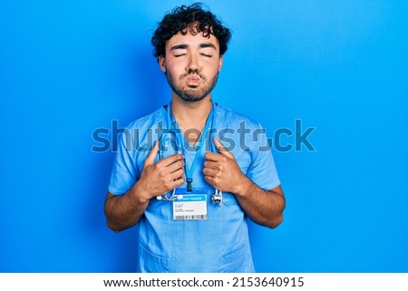 Young hispanic man wearing blue male nurse uniform puffing cheeks with funny face. mouth inflated with air, catching air. 