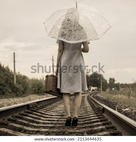 Vintage style. A girl in a dress and heels with an umbrella and a suitcase goes by rail on foot. Rear view. No face. A retro-style girl walking off into the distance by rail. Royalty-Free Stock Photo #2153644635