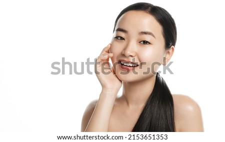 Beautiful asian woman with hydrated, moisturized skin, touches her glowing face and smiles at camera, stands with bare shoulders, white background