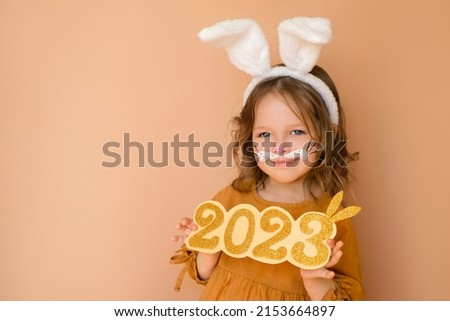 Kid with painted face in guise of rabbit holds number of new year 2023. Zodiac sign according to Eastern Chinese calendar. Cute person dressed up with bunny ears and makeup in anticipation of new year Royalty-Free Stock Photo #2153664897