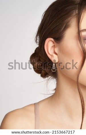 Cropped side portrait of a brown-haired lady in golden round stud earrings with a pendant made as an incandescent light bulb. The girl is posing on the gray background. Royalty-Free Stock Photo #2153667217