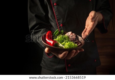 Preparation of beef steak and fresh vegetables. Plate with food in the chef hand. Dark background free space for advertising.