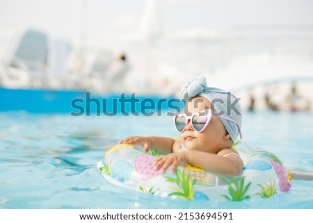 Cute funny toddler girl in colorful swimsuit and sunglasses relaxing on inflatable toy ring floating in pool have fun during summer vacation in tropical resort. Child having fun in swimming pool.  Royalty-Free Stock Photo #2153694591