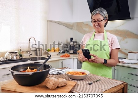 Adult woman, cooking a Spanish dish, estofado de albóndigas. Taking a cell phone photo of the finished meal for social media. Wearing an apron, wearing glasses and cooking utensils.