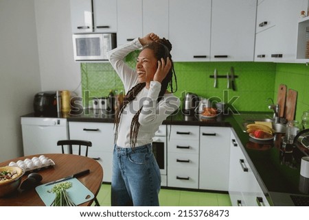 Positive young woman dancing and singing while listening to music in headphones in kitchen