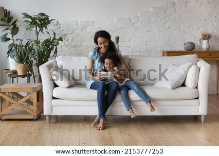 Happy Black mom and cheerful preschool daughter girl reading book online on tablet, using online learning app on digital gadget, resting on couch, enjoying leisure in stylish eco home interior