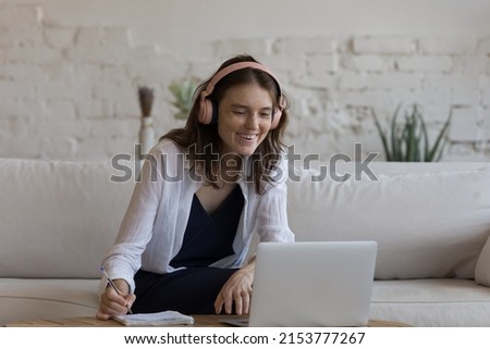 Happy focused gen Z student girl in big wireless headset watching webinar, online tutorials on laptop, studying language with online class, speaking to teacher on video call, writing notes, smiling