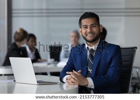Happy cheerful Indian professional guy head shot portrait at laptop in co-working space with diverse business group in background. Businessman working at rental workplace, looking at camera Royalty-Free Stock Photo #2153777305