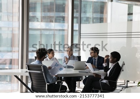 Serious adult female business leader instructing diverse team on brainstorming session at meeting table. Group of coworkers, employees discussing paper marketing reports, project analytics Royalty-Free Stock Photo #2153777341