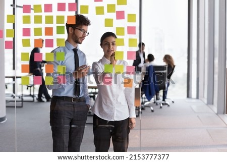 Confident Indian mentor woman teaching young Intern guy, training to use Kanban board for planning tasks, project strategy. Diverse millennial employees using scrum management for teamwork Royalty-Free Stock Photo #2153777377