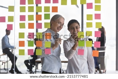 Happy senior business coach and young Indian employee studying scrum management, writing notes on stickers on glass board, planning tasks, working on project strategy, smiling, laughing