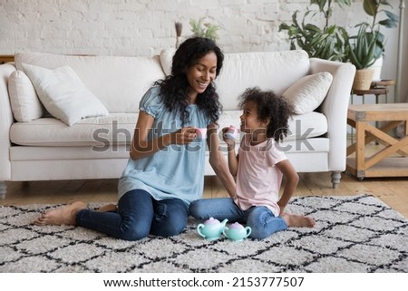 Cheerful Black young mother and little daughter kid having fun, playing tea party, pretending drinking coffee from toy cups, playing funny role games on warm heating floor in cozy living room Royalty-Free Stock Photo #2153777507