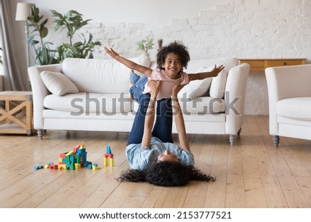 Happy carefree little Black kid playing airplane with mom, looking at camera, smiling. Mother lifting daughter child with flying hands in air, resting on heating floor, exercising with excited girl Royalty-Free Stock Photo #2153777521