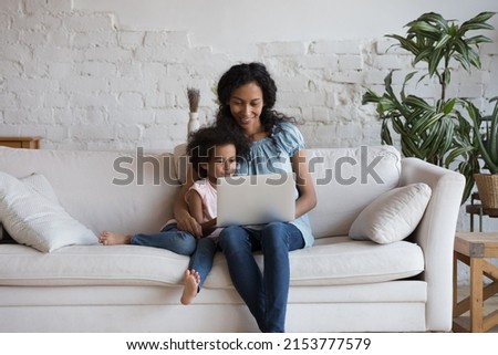 Curious adorable daughter girl and happy African mom reading online book, using learning app on laptop, watching cartoon movie, series, talking on video call, shopping. Family communication
