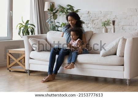 African American mom teaching little daughter girl to use learning app on laptop. Mother and kid watching cartoon movie together, enjoying leisure in living room eco interior