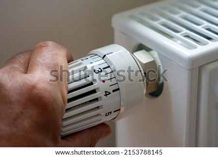 Rising heating costs in the crisis: Man regulates the temperature at home with the heating thermostat to save energy, close up with hand Royalty-Free Stock Photo #2153788145