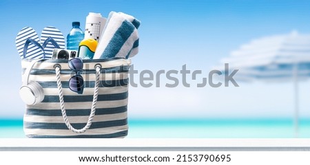 Stylish beach bag with accessories and tropical beach in the background, summer vacations concept