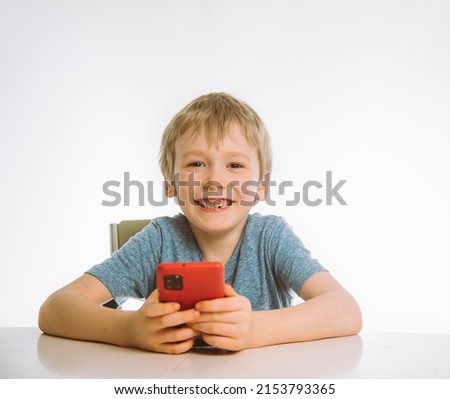the boy is sitting at the table holding a smartphone and smiling, white background. copy space.