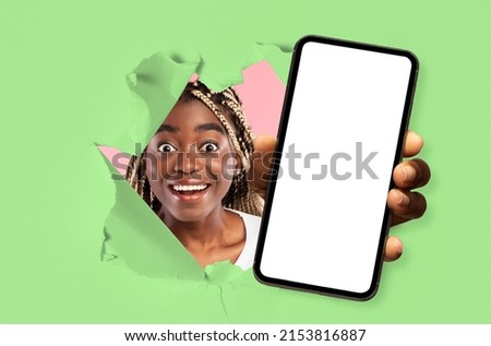 Wow, Great Offer. Excited young black lady holding big smartphone with white blank screen in hand, showing close up to camera through torn green paper hole. Gadget with empty free copy space, banner