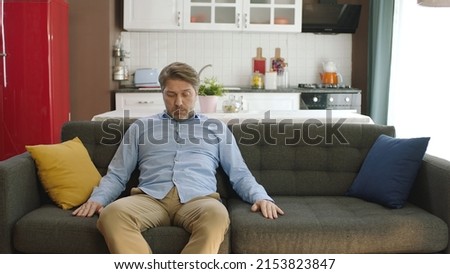 Thoughtful young man sitting alone on sofa at home. Bored and pensive male sitting at home alone thoughtfully in his chair. Uncertainty concept.