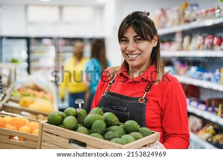 Happy latin woman working inside supermarket while holding wood box with avocados Royalty-Free Stock Photo #2153824969