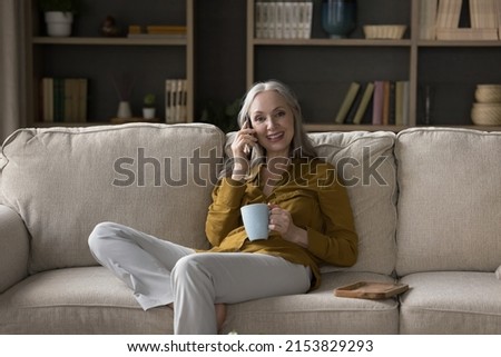 Happy pretty mature woman speaking on mobile phone portrait. Senior grey haired lady making telephone call, sitting on couch at home, enjoying talk, communication, looking at camera, smiling