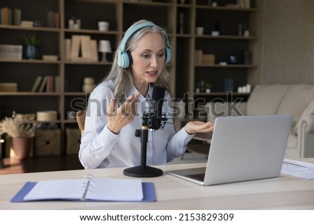 Elder grey haired teacher woman using laptop, wireless headphones, professional microphone, giving webinar, lecture, recording learning podcast holding online class, teaching audience of students