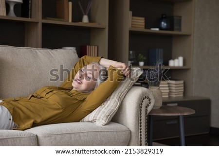 Carefree happy retired woman enjoying break, leisure time at home, relaxing, lying on comfortable couch, smiling, laughing, looking away, breathing fresh air. Stress relief concept Royalty-Free Stock Photo #2153829319