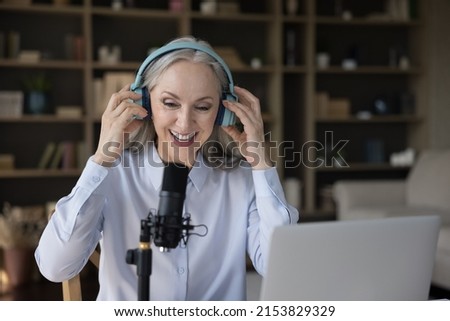 Mature blogger woman in headphones speaking at professional microphone at laptop, taking video for blog. Senior radio host, speaker holding program on air, broadcasting, recording audio book Royalty-Free Stock Photo #2153829329