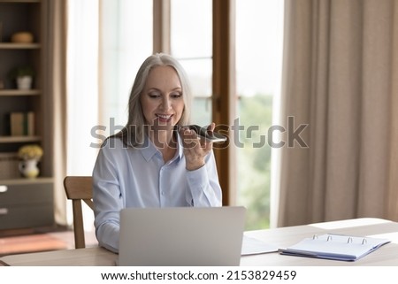 Happy grey haired freelance business lady using virtual assistant on mobile phone, giving voice command, recording audio message on smartphone at laptop computer, working in home office,