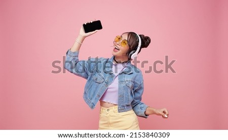 Happy beautiful Asian teen woman in yellow glasses listening music in headphones and dance on pink background.