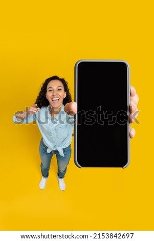 Cheerful young woman holding cell phone in hand pointing at gadget with blank black mockup screen smiling on yellow orange wall, recommending newest mobile app, above high angle top view, copy space Royalty-Free Stock Photo #2153842697