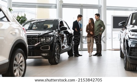 Car purchase or rental. Positive young spouses speaking to salesman about buying new auto at dealership, panorama. Successful millennial manager showing clients choice of vehicles at salon Royalty-Free Stock Photo #2153842705