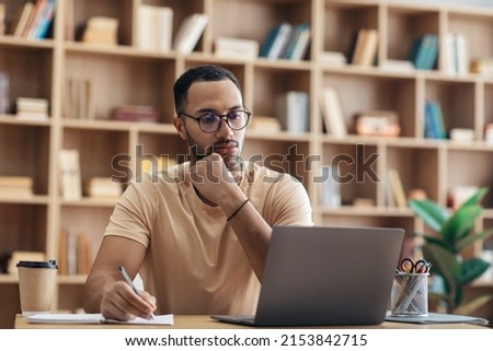 Focused arab man in glasses using laptop sitting at desk and writing in notebook, concentrated guy taking notes, watching webinar studying online, free space
