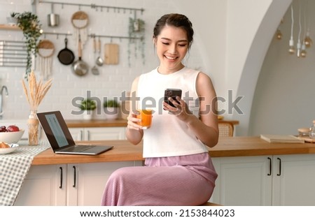 Young woman Asian happy rest drinking orange juice and using mobile phone in kitchen room.