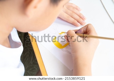 Close up of cute Asian little boy drawing and painting his imagination monster character on white art paper at home. Child brain development and art therapy concept.