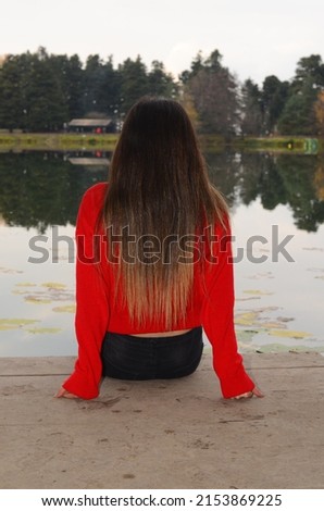 Girl with long hair sitting by the lake. Portrait of beautiful girl with long hair wearing red color sweater. Symbol of loneliness. selective focus.
 Royalty-Free Stock Photo #2153869225