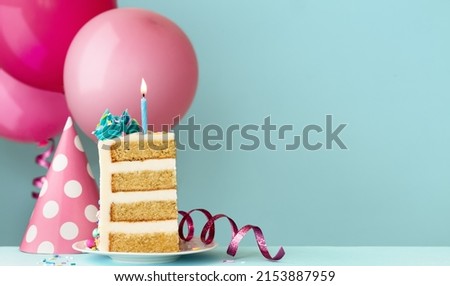 Slice of birthday cake with blue birthday candle, party hat, streamers and birthday balloons ready for a birthday party Royalty-Free Stock Photo #2153887959