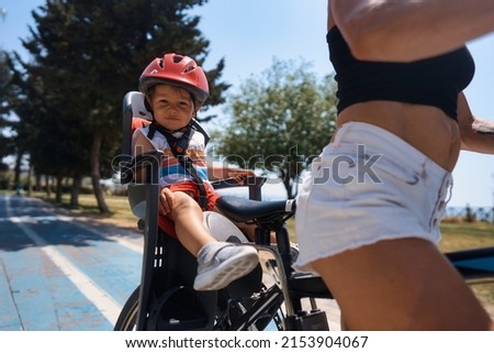 Portrait of little toddler boy with security helmet on the head sitting in bike seat and his mother with bicycle. Safe and child protection concept. Family and weekend activity trip Royalty-Free Stock Photo #2153904067