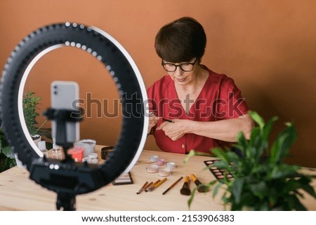 Middle-aged woman talking on cosmetics with makeup eye shadows and blush palette and brushes while recording her video. Mature female making video for her blog on cosmetics