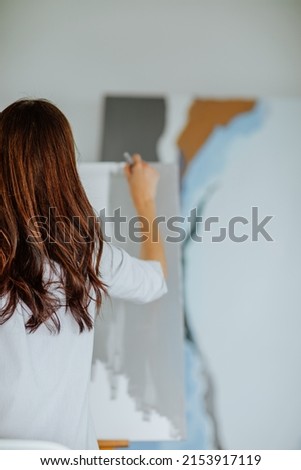 Young brunette woman in blue shirt painting big grey picture stands on the easel at home. View from the back. Art concept.