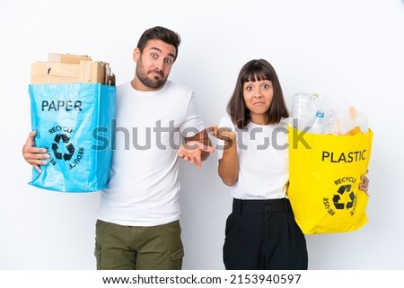 Young couple holding a bag full of plastic and paper to recycle isolated on white background having doubts while raising hands and shoulders