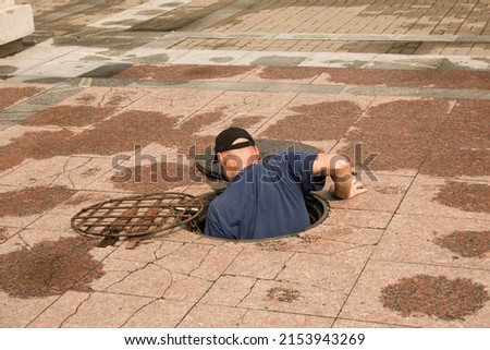 A worker climbs out of an open manhole in the road. Dangerous open unprotected manhole on the road. Accident with a sewer manhole in the city. The concept of repairing underground utilities. Royalty-Free Stock Photo #2153943269