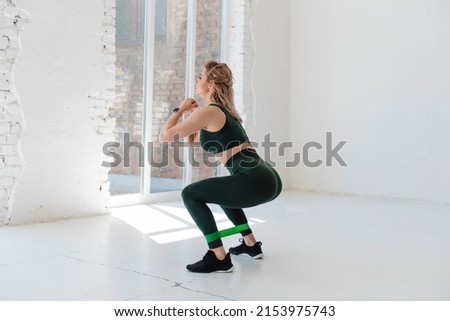 Side view of fitness young woman in green sportswear using rubber resistance band for legs exercises. Attractive girl with blond hair doing stretching domestic workout.