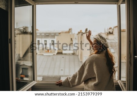 A woman stands near the window against the backdrop of the roofs of Paris. A girl with long hair in a beige suit and a beret enjoys the city and the trip waving to her neighbors. Royalty-Free Stock Photo #2153979399