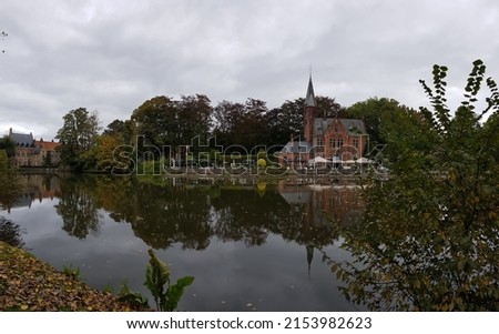 Panoramic view of a pond with reflection, red building and café on the other side and a few more houses. Trees, bush, yellow, green, autumn. On this side yellow leaves, trees. Brugge, Belgium,Oct 2021 Royalty-Free Stock Photo #2153982623