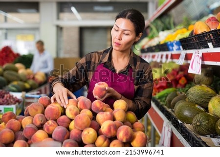 Asian woman in apron stacking peaches in salesroom of greengrocer.