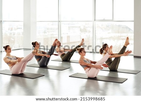 Group of young women sitting on yoga mats and doing boat pose in modern studio with panoramic windows Royalty-Free Stock Photo #2153998685