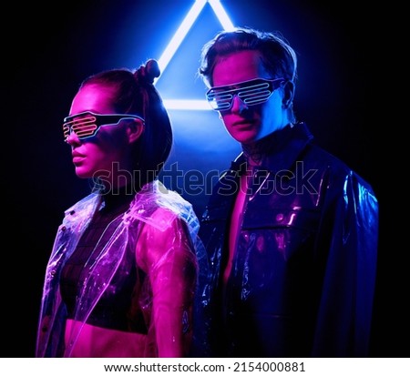 Portrait of confident young people in fashionable cyber outfits standing in dark room with neon triangle sign
