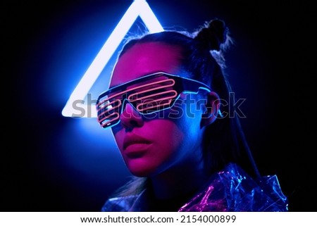Portrait of serious super girl in neon flashing goggles standing in dark room with neon illumination Royalty-Free Stock Photo #2154000899